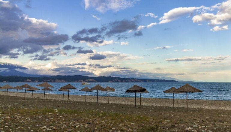 Абхазия в июне 2021 _Straw beach umbrellas on the Empty Black Sea coast in Abkhazia, Pitsunda at sunset. Pebble beach, mountains, sea, sky with clouds