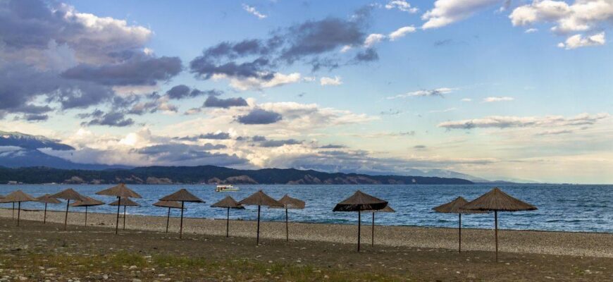 Абхазия в июне 2021 _Straw beach umbrellas on the Empty Black Sea coast in Abkhazia, Pitsunda at sunset. Pebble beach, mountains, sea, sky with clouds